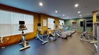 The 56 Broad Street Fitness Center in Bloomfield, NJ, is a well-equipped, single-floor facility designed to support a full range of workout routines. The space is accessed through a secure entry vestibule featuring both a solid door and glass double doors, with a hand sanitizer dispenser and keycard access for user convenience.

The main workout area is divided into two distinct zones. The cardio section is lined with multiple treadmills and elliptical trainers, each equipped with individual monitors, as well as upright and recumbent stationary bikes and stair steppers. Large windows with white blinds allow natural light to fill this area, complementing the recessed ceiling lighting and warm amber-toned walls.

The strength training zone features a robust selection of cable machines, multi-use weight stations, and functional training equipment. A striking faux-green wall accent adds a dynamic visual element, while large wall-mounted mirrors amplify the sense of space. Exercise accessories including stability balls, medicine balls, and foam rollers are neatly organized on dedicated racks. A wall-mounted flat-screen TV provides entertainment throughout the space.

The floors throughout are finished with durable, dark speckled rubber flooring suited for high-traffic athletic use. The overall ambiance is energetic yet functional, offering a comprehensive fitness environment for all levels of users.
