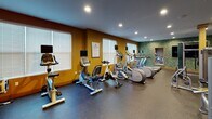 The 56 Broad Street Fitness Center in Bloomfield, NJ, is a well-equipped, single-floor facility designed to support a full range of workout routines. The space is accessed through a secure entry vestibule featuring both a solid door and glass double doors, with a hand sanitizer dispenser and keycard access for user convenience.

The main workout area is divided into two distinct zones. The cardio section is lined with multiple treadmills and elliptical trainers, each equipped with individual monitors, as well as upright and recumbent stationary bikes and stair steppers. Large windows with white blinds allow natural light to fill this area, complementing the recessed ceiling lighting and warm amber-toned walls.

The strength training zone features a robust selection of cable machines, multi-use weight stations, and functional training equipment. A striking faux-green wall accent adds a dynamic visual element, while large wall-mounted mirrors amplify the sense of space. Exercise accessories including stability balls, medicine balls, and foam rollers are neatly organized on dedicated racks. A wall-mounted flat-screen TV provides entertainment throughout the space.

The floors throughout are finished with durable, dark speckled rubber flooring suited for high-traffic athletic use. The overall ambiance is energetic yet functional, offering a comprehensive fitness environment for all levels of users.