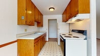 This single-floor residential space presents a practical and classic layout designed for comfortable everyday living. The galley-style kitchen is efficiently organized with warm honey-toned wood cabinetry lining both walls, providing generous storage from floor to ceiling. Light-colored countertops offer ample workspace, while white appliances — including an electric range and refrigerator — complement the patterned vinyl tile flooring. A range hood and flush-mount ceiling light complete the functional setup, with a doorway at the end leading further into the unit.

The living area is generously proportioned, featuring plush wall-to-wall carpeting, crisp white walls, and elegant crown molding that lends a subtle refined touch. A large sliding glass door floods the room with natural light and provides outdoor access, while a baseboard heating system runs along the perimeter. An adjacent carpeted bedroom also benefits from natural light through a framed window with views to the exterior.

The bathroom is well-appointed with warm wood vanity cabinetry, a wide mirror with overhead lighting, mosaic tile flooring, and full white wall tile surrounding the tub and shower area. A second bathroom features classic yellow and brown ceramic wall tiles with a grab bar, adding a retro character. Throughout, the space maintains a clean, neutral palette with consistent wood trim detailing.