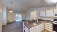 This single-floor residence offers a thoughtfully designed open-concept layout that seamlessly connects the kitchen and living areas. The space is distinguished by striking vaulted ceilings that lend an airy, expansive feel throughout the main living area. Large windows and a sliding glass door leading to a private balcony flood the interior with natural light and provide pleasant outdoor views.

The kitchen is efficiently laid out in an L-shape and features white cabinetry with warm wood-tone trim, dark laminate countertops, a double-basin sink, a full suite of appliances including a refrigerator, electric range, built-in microwave, and a dishwasher. A peninsula counter provides a natural divide between the kitchen and the open living space.

The carpeted living and dining areas benefit from the soaring vaulted ceiling, with a pendant light fixture adding a decorative touch. A standout architectural detail is a deeply recessed picture window with built-in shelving beneath it, offering both visual interest and functional display space. The bathroom features a wide vanity with ample cabinetry, a large mirror, and well-placed lighting. An in-unit laundry area is also present. Throughout, the neutral color palette and clean finishes create a bright, welcoming ambiance.
