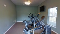 The Jefferson Post fitness center offers a dedicated, single-floor workout facility designed to meet residents' exercise needs. The room is finished with cool sage-green walls and carpeted flooring, creating a calm and focused environment for physical activity. A ceiling fan with an integrated light fixture provides both ventilation and illumination, keeping the space comfortable during workouts. A wall-mounted flat-screen television is positioned at an ideal viewing height, allowing residents to stay entertained while exercising. The room is well-equipped with multiple cardio machines, including ellipticals and additional aerobic equipment, arranged to maximize usable floor space. Electrical outlets and ventilation are thoughtfully placed throughout the room to ensure convenience and airflow. The layout is open and functional, making it easy to move between machines. This fitness space reflects a commitment to providing residents with accessible, on-site amenities that support an active lifestyle within the Jefferson Post community.