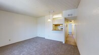 This single-floor space presents an open-concept layout connecting the living area to the kitchen. The living room is finished with wall-to-wall carpeting in a neutral gray-beige tone and features a textured popcorn ceiling. A pendant light fixture hangs near the kitchen pass-through, adding a focal point to the space. The kitchen is partially visible through a raised bar-style pass-through counter, offering wood-toned cabinetry, a fluorescent overhead light, and white appliances. The layout encourages a natural flow between the living and kitchen areas. From the living room, a hallway extends toward additional rooms, illuminated by overhead lighting and finished with hard-surface flooring, contrasting the carpeted main area. The walls throughout are painted in a clean, bright white, contributing to a light and airy feel. HVAC venting is visible near the ceiling, indicating a central air system. Overall, the space presents a functional, straightforward layout with neutral finishes designed to complement a variety of furnishing styles.