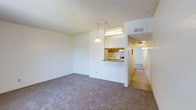 This single-floor space presents an open-concept layout connecting the living area to the kitchen. The living room is finished with wall-to-wall carpeting in a neutral gray-beige tone and features a textured popcorn ceiling. A pendant light fixture hangs near the kitchen pass-through, adding a focal point to the space. The kitchen is partially visible through a raised bar-style pass-through counter, offering wood-toned cabinetry, a fluorescent overhead light, and white appliances. The layout encourages a natural flow between the living and kitchen areas. From the living room, a hallway extends toward additional rooms, illuminated by overhead lighting and finished with hard-surface flooring, contrasting the carpeted main area. The walls throughout are painted in a clean, bright white, contributing to a light and airy feel. HVAC venting is visible near the ceiling, indicating a central air system. Overall, the space presents a functional, straightforward layout with neutral finishes designed to complement a variety of furnishing styles.