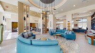 This contemporary amenity facility is thoughtfully designed to offer residents a comprehensive lifestyle experience across multiple distinct spaces. The expansive fitness center is divided into dedicated zones, including a cardio area outfitted with treadmills, ellipticals, and stationary bikes with personal screens, a strength training zone with cable machines and free weights, and a separate functional training studio featuring a RAX rack system, medicine balls, BOSU trainers, and a roll-up glass garage door that opens to the outdoors. Vibrant geometric murals and full-length mirrors energize the workout environment, while exposed blue ductwork adds an industrial accent beneath soaring ceilings. The social lounge areas are equally impressive, anchored by a linear fireplace set against a floor-to-ceiling textured tile surround, flanked by illuminated built-in shelving. Comfortable teal seating arrangements and warm wood accents create an inviting atmosphere. A bright communal dining and game area features terrazzo flooring, dramatic copper pendant lighting, clerestory windows, and direct access to an outdoor terrace. A stylish kitchenette bar with a bold Joshua tree mural serves as a striking focal point, complemented by globe pendant lights and yellow upholstered barstools. Throughout, the design blends modern aesthetics with warm, livable details.