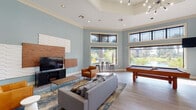 Located in Humble, TX, this well-appointed clubroom is designed to deliver a comfortable and social atmosphere for its users. The space features soaring ceilings with elegant crown molding and a tray ceiling detail, lending an airy, upscale feel throughout. Soft blue-gray walls complement light wood-style flooring, creating a cohesive and modern aesthetic.

The main lounge area is furnished with a plush gray sectional sofa, rich cognac leather armchairs, and a wood-and-glass coffee table arranged around a large flat-screen TV mounted on a striking mixed-material accent wall featuring textured white panels and warm wood elements. Adjacent to the lounge, a full-size pool table provides an engaging recreational option.

High, large-pane windows line the perimeter of the room, flooding the interior with abundant natural light and offering picturesque views of lush landscaping, palm trees, and an outdoor pool area. High transom windows add to the sense of openness and height.

A dedicated kitchen area features rich dark wood cabinetry, dark countertops, and black appliances, including a refrigerator and gas range. A spacious bar-height counter with round leather stools provides casual seating for dining or socializing. Additional bistro-style table seating near the windows offers a bright spot for informal gatherings, completing this versatile and inviting community space.