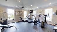 Located at 1600 Belle Point Dr in Mt. Pleasant, South Carolina, this dedicated fitness center offers a clean, well-organized workout environment designed to meet a variety of exercise needs. The single-floor space features durable rubber flooring throughout, providing both safety and comfort during workouts. The room is outfitted with multiple treadmills, an elliptical trainer, a stationary bike, and additional cardio equipment, making it a comprehensive facility for cardiovascular exercise. A wall-mounted flat-screen television is positioned for easy viewing during workouts, adding a convenient entertainment element to the space. Natural light floods the room through multiple windows along the walls, creating a bright and energizing atmosphere. Recessed lighting and a ceiling fan with integrated light ensure the space remains well-lit and comfortable at any time of day. Crown molding along the ceiling adds a refined architectural touch to the otherwise functional space. Informational charts and maps are displayed on the walls, offering useful references for fitness routines. The overall ambiance is clean, open, and welcoming, reflecting a commitment to providing a quality fitness experience in a comfortable community setting.