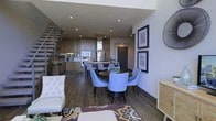 This contemporary two-story residence showcases a thoughtfully designed open-concept main floor that seamlessly integrates the kitchen, dining, and living areas into a cohesive and functional layout. The kitchen is appointed with rich wood cabinetry, stainless steel appliances including a built-in wall oven, and a central island that serves as both a prep space and a casual gathering point. Recessed lighting throughout the ceiling enhances the modern aesthetic while keeping the interior bright and inviting. The dining area features a round dark-wood table surrounded by upholstered chairs in a soft blue-grey tone, creating an elegant yet approachable setting for meals and entertaining. Warm hardwood flooring runs throughout the main level, adding depth and continuity to the space. The living area is anchored by a tufted accent chair and a colorful area rug that introduces a vibrant focal point. A striking open-tread staircase with metal railings connects the two floors, adding an architectural element that is both functional and visually compelling. Framed wall art and carefully selected accent pieces contribute to the polished, curated character of the home.