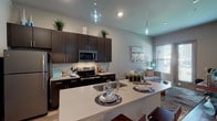 Located in Twin Oaks, MO, this single-floor residence showcases a thoughtfully designed open-concept layout with modern finishes throughout. The kitchen is a standout feature, equipped with dark espresso cabinetry, stainless steel appliances including a refrigerator, microwave, and range, complemented by a tiled backsplash and a large quartz-topped island with an undermount sink and bar seating. Stylish pendant lighting hangs above the island, adding a contemporary touch. The kitchen flows seamlessly into the living area, where natural light pours through large windows and a glass-paneled door leading to an outdoor view. The living space is accented by elevated ceilings that enhance the sense of openness. Two bedrooms provide comfortable accommodations, each featuring carpeted flooring, generous windows, and ample natural light. A walk-in closet with wire shelving connects to a full bathroom with a shower-tub combo. A second bathroom features a single-sink vanity with dark cabinetry, a large mirror, and wood-look flooring. Throughout the residence, light hardwood-style floors in the main areas and neutral gray tones on the walls create a cohesive, contemporary aesthetic that is both functional and visually appealing.