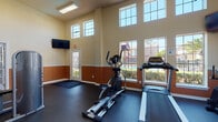 This fitness center is designed to offer a bright and motivating workout environment. The space is distinguished by its impressively tall ceilings, which lend an open, airy feel throughout. A two-tone wall treatment — warm beige on the upper half and a rich terracotta orange on the lower half — gives the room a distinctive and energetic aesthetic. Durable rubber flooring covers the entire workout area, providing both safety and comfort underfoot.

Natural light pours in through an array of large, grid-pane windows positioned along the walls and at clerestory height, ensuring the space remains well-lit throughout the day. Direct access to the outdoor pool area is provided through a glass-paneled door, offering pleasant views of the surrounding grounds and greenery.

The fitness center is equipped with a variety of cardio machines, including treadmills and an elliptical trainer, positioned to take advantage of the outdoor views. Strength training is supported by multiple cable and weight machines, including an Inflight Fitness seated machine and a lat pulldown/low row station. A wall-mounted flat-screen television is positioned for easy viewing during workouts. The space connects to an adjacent lounge or common area, adding convenience for users before or after their exercise sessions.
