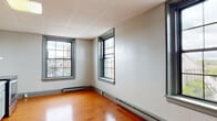 Situated at 35 Union St in New London, Connecticut, this single-floor unit offers a practical and functional layout with a clean, understated aesthetic throughout. The main living area is characterized by warm, rich hardwood flooring that carries consistently through the space, paired with neutral white walls and a drop-panel ceiling. Multiple large, grid-paned windows allow natural light to filter in while offering elevated views of the surrounding New London streetscape and cityscape, providing a pleasant urban backdrop. Baseboard heating runs along the perimeter of the rooms, ensuring year-round comfort. The kitchen is efficiently designed, featuring soft gray cabinetry with decorative raised-panel doors, a granite-style countertop, a stainless steel refrigerator, a stainless steel electric range, and a tile backsplash that adds a subtle textural element. A range hood is positioned above the stove for ventilation. The open-concept connection between the kitchen and living area contributes to an airy, unobstructed flow. The bathroom includes tile flooring, a toilet, and an accessible grab bar, reflecting a straightforward and functional design. Overall, the space presents a no-frills, move-in-ready environment well-suited for those seeking a practical urban residence in downtown New London.