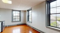 Situated at 35 Union St in New London, Connecticut, this single-floor unit offers a practical and functional layout with a clean, understated aesthetic throughout. The main living area is characterized by warm, rich hardwood flooring that carries consistently through the space, paired with neutral white walls and a drop-panel ceiling. Multiple large, grid-paned windows allow natural light to filter in while offering elevated views of the surrounding New London streetscape and cityscape, providing a pleasant urban backdrop. Baseboard heating runs along the perimeter of the rooms, ensuring year-round comfort. The kitchen is efficiently designed, featuring soft gray cabinetry with decorative raised-panel doors, a granite-style countertop, a stainless steel refrigerator, a stainless steel electric range, and a tile backsplash that adds a subtle textural element. A range hood is positioned above the stove for ventilation. The open-concept connection between the kitchen and living area contributes to an airy, unobstructed flow. The bathroom includes tile flooring, a toilet, and an accessible grab bar, reflecting a straightforward and functional design. Overall, the space presents a no-frills, move-in-ready environment well-suited for those seeking a practical urban residence in downtown New London.