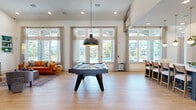 This generously sized single-floor community clubhouse is thoughtfully designed to accommodate a variety of social and functional activities. The entrance foyer makes an immediate impression with soaring ceilings, a textured accent wall, bold artwork, and warm wood-look tile flooring that carries throughout the space.

The main gathering area is anchored by a full-size pool table beneath a striking industrial pendant light, surrounded by floor-to-ceiling windows that flood the room with natural light and offer views of an outdoor pool area. A vaulted ceiling with teal accent walls and built-in shelving adds architectural character to this zone, which also includes a comfortable lounge with seating and a wall-mounted television.

Adjacent to the game and lounge area, a sleek bar and kitchen features navy cabinetry, white quartz countertops, stainless steel appliances, and a long communal counter with bar seating illuminated by gold globe pendant lights.

A glass-paneled barn door separates the business center, which offers a quieter setting equipped with a workstation, built-in cabinetry, carpeted flooring, and direct exterior access. The overall ambiance balances modern design with welcoming warmth throughout.