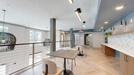 Located at 3902 W Tyvola Rd in Charlotte, NC, this single-floor common area serves as a welcoming lobby and amenity space with a cohesive modern design throughout. The open-plan layout is anchored by light-toned herringbone-patterned flooring that lends a refined, geometric quality to the space.

A prominent reception counter runs along one wall, finished with warm wood cabinetry, a crisp white quartz countertop, and a bold geometric patterned tile backsplash in white and grey. Brass wall sconces and recessed lighting illuminate the counter area, while potted greenery adds a natural accent. Built into the cabinetry is a beverage refrigerator and a stainless steel sink, equipping the counter for functional use.

The lounge area features a mix of casual seating around a wood-topped dining table, beneath sculptural woven pendant lights that hang from the elevated ceiling. Dark structural columns frame the space and provide visual contrast against the light walls and floors. A cable-railing mezzanine overlooks a lower level, with large windows bringing in natural light and exterior views of the surrounding greenery. Stainless steel elevator doors are conveniently located nearby, completing this well-appointed and functional communal space.