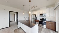 This well-appointed single-floor unit showcases a thoughtfully designed open-concept layout that seamlessly connects the kitchen and living areas. The kitchen serves as a central focal point, anchored by a large island with dark granite countertops, an integrated sink with a brushed gold faucet, and dual pendant lights overhead. White upper cabinetry pairs with rich wood-toned lower cabinets, complemented by a classic subway tile backsplash and a full suite of stainless steel appliances, including a refrigerator, range, and microwave. Track lighting above provides additional illumination throughout the space. The adjacent living area is generously proportioned, featuring plush carpeting and large windows that invite abundant natural light and offer views of the surrounding neighborhood. A sliding glass door opens to a private balcony, extending the living space outdoors. The bedroom is a serene retreat with soft carpeting, dual windows framing neighborhood views, and direct access to a walk-in closet. The bathroom features a soaking tub with white subway tile surround, wood-look flooring, and a granite-topped vanity with warm wood cabinetry. A dedicated laundry area with a full-size washer and dryer and built-in shelving completes this functional and stylish home.