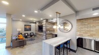 This single-floor amenity lounge is designed with a contemporary aesthetic that blends industrial and warm natural elements throughout. The entry corridor features floor-to-ceiling glass partitions with sleek dark frames, offering clear sightlines into the space while maintaining a polished, modern feel. Large-format stone tile flooring flows consistently throughout, anchoring the open layout.

The lounge is divided into distinct yet connected zones. A comfortable seating area includes a leather sofa with bold orange accent pillows, upholstered arm chairs, and a patterned area rug beneath a substantial drum pendant light. Tall windows admit natural light and provide views to the exterior. A striking reclaimed wood accent wall serves as a focal point, housing a large flat-screen television and flanked by additional seating for media viewing.

Adjacent to the lounge is a well-equipped wet bar area featuring yellow brick tile backsplash, dark cabinetry, a sink, and stainless steel appliances. A freestanding marble-topped bar with blue upholstered stools is suspended beneath a multi-globe brass pendant light and a decorative circular mirror. A separate nook with exposed brick walls displays bold abstract artwork above two rounded lounge chairs, offering a quieter retreat within the broader space.