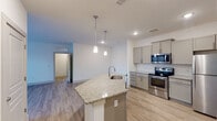 This well-appointed single-floor residence offers a clean, contemporary design throughout. The open-concept kitchen serves as the centerpiece of the living area, anchored by a spacious island with a granite countertop and an integrated sink, illuminated by stylish pendant lighting. Gray shaker-style cabinetry lines the walls, complemented by a classic white subway tile backsplash and a full suite of stainless steel appliances, including an electric range, over-the-range microwave, and refrigerator. Light wood-style flooring flows through the main living and kitchen areas, creating a bright, cohesive aesthetic. The living area is open and airy, with recessed lighting and a door leading to an outdoor space, allowing for natural light to filter in. A hallway connects the main living area to the private quarters, which feature neutral carpet flooring and crisp white walls. The bathroom is finished with a granite-topped vanity with a rectangular undermount sink, a large mirror, and a full tub-shower combo with white tile surround. Consistent granite countertop finishes and gray cabinetry tie the design together cohesively throughout the space, offering a polished and functional living environment.