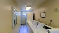 This shared laundry facility is a functional, well-organized space designed to accommodate multiple users efficiently. The room features a row of top-loading washing machines lined along one side, complemented by stacked dryer units positioned toward the back of the space. A continuous white countertop surface runs alongside the washers, providing a convenient area for sorting and folding laundry. The flooring consists of light-colored tiles that are easy to maintain and contribute to the clean, utilitarian aesthetic of the room. Overhead, flush-mounted fluorescent light fixtures provide bright, even illumination throughout the space, while a small window near the rear allows a measure of natural light to filter in. The walls are finished in a neutral beige tone, lending the room a simple and practical atmosphere. A glass-front bulletin or display case is mounted on one wall near the entrance, likely used for posting notices or instructions, and a framed informational sign is visible on the adjacent wall. The layout is straightforward and efficient, allowing comfortable movement between machines. Overall, the facility is clean, well-lit, and thoughtfully arranged to serve its purpose effectively.