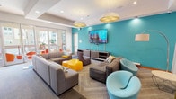 This single-floor amenity space is designed as a dynamic social hub, combining a comfortable lounge area with an active game room. The lounge is anchored by a bold teal accent wall that serves as a backdrop for a large wall-mounted flat-screen TV, complemented by a wood media console with open shelving. Seating is plentiful and eclectic, featuring modular gray sofas, a deep brown sectional, a sculptural lavender chair, and playful light blue swivel chairs — all arranged to encourage conversation and group viewing. Vibrant yellow hexagonal ottomans add a pop of color to the center of the seating arrangement. Overhead, distinctive gold-toned hexagonal pendant lights and recessed ceiling fixtures provide warm, layered illumination, while a coffered ceiling detail adds architectural interest. Floor-to-ceiling glass doors open to an outdoor balcony area furnished with bright orange seating, allowing ample natural light to pour into the space. Adjacent to the lounge, the game room features a full-size pool table, a foosball table, and dual wall-mounted screens against a bold yellow accent wall, offering a dedicated area for active recreation. Patterned carpet flooring ties the two zones together cohesively, creating an inviting and energetic atmosphere throughout.