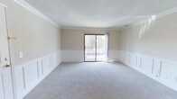 This single-floor space is thoughtfully designed with classic architectural details that lend it a polished, refined character. The main living area is a standout feature, showcasing crisp white wainscoting panels that line the lower half of the walls, adding a timeless elegance to the room. Above the wainscoting, the walls are finished in a soft, warm neutral tone that creates a calm and inviting atmosphere throughout.

The room is finished with crown molding that runs along the ceiling perimeter, further enhancing the traditional aesthetic of the space. Plush, light gray carpeting covers the floor, providing a comfortable and cohesive foundation underfoot.

Natural light is a defining characteristic of this space, flowing generously through a large sliding glass door that serves as both a focal point and a functional access point to an outdoor balcony. The balcony is enclosed with a classic railing and overlooks a lush, tree-lined setting, offering a pleasant connection to the outdoors. The textured ceiling and overall neutral palette work together to create a bright, airy, and welcoming environment throughout this well-appointed single-floor space.