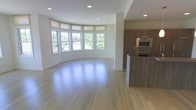 This single-floor space showcases an open-concept layout that seamlessly connects the living area and kitchen. The living area is defined by a striking curved bay window wall, which floods the room with abundant natural light and offers views of lush greenery outside. Recessed ceiling lighting complements the natural light, keeping the space bright and airy throughout. Light-toned hardwood flooring extends across the entire space, reflecting light and contributing to the clean, modern aesthetic.

The kitchen is positioned to one side and features warm, medium-toned wood cabinetry that provides a natural contrast to the white walls. Stainless steel appliances, including a built-in wall oven and microwave, are seamlessly integrated into the cabinetry for a polished, contemporary look. A kitchen island with matching wood paneling adds functional workspace and subtly delineates the kitchen from the living area without disrupting the open flow.

The overall ambiance is bright, modern, and welcoming, with a neutral color palette of whites, warm wood tones, and light floors creating a versatile and elegant interior.