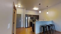 This single-floor space showcases a well-appointed kitchen as its central feature. Dark espresso-toned cabinetry lines the walls, providing ample storage and creating a bold contrast against the light-colored walls and warm hardwood-style flooring that runs throughout. The kitchen is equipped with stainless steel appliances, including a full-size refrigerator, a built-in microwave, and a range, all contributing to a cohesive, modern aesthetic. A peninsula with a granite-style countertop extends into the living area, doubling as a breakfast bar with industrial-style metal stools tucked neatly underneath. Pendant lights hang above the bar, adding focused ambient lighting that complements the flush-mount ceiling fixtures. The ceilings are noticeably elevated, lending an open, airy quality to the space. A tiled backsplash adds a subtle decorative touch behind the range. Near the entryway, a solid paneled door with a deadbolt provides a secure and defined entry point. Overall, the space maintains a clean, contemporary feel with a practical layout designed for comfortable everyday living.