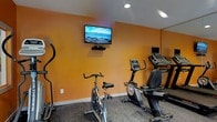 This single-floor fitness space is thoughtfully designed to provide a dedicated environment for cardiovascular exercise and overall wellness. The room is defined by its warm, vibrant orange accent walls, which lend an energizing ambiance well-suited to an active workout setting. Recessed ceiling lighting ensures the space is well-lit, creating a bright and focused atmosphere for exercise.

The room is outfitted with a selection of cardio equipment, including a stationary spin bike, a recumbent bike, and a treadmill, offering a variety of workout options to suit different fitness preferences and goals. An additional piece of cardio equipment, resembling an elliptical or stepper, rounds out the available machines. A wall-mounted flat-screen television is positioned at eye level, providing entertainment during workouts, while a secondary monitor is also visible near the treadmill area for added convenience.

The flooring features a durable, dark-toned carpet suited to a fitness environment. The overall layout is efficient and practical, making the most of the available space while keeping all equipment easily accessible. This fitness room presents a functional and motivating setting for maintaining an active routine.