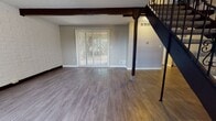 This two-story space is defined by its open, airy main level, where hardwood-style flooring extends throughout the living areas and a distinctive dark wood exposed beam runs across the ceiling, adding architectural character. A bold staircase with ornamental wrought iron railings serves as a central design feature, connecting both floors while contributing a classic, elegant aesthetic to the space.

The main level offers a generous living area illuminated by natural light from vertical blind-covered sliding glass doors and windows that frame views of surrounding greenery. A dedicated entryway area provides a welcoming transition into the home. The kitchen is equipped with wood cabinetry, white appliances including an electric range and dishwasher, and hard-surface flooring, with a window allowing natural light. A half bathroom on the main level features a wood vanity with mirrored cabinet above and tile flooring.

The upper level hosts multiple bedrooms, each carpeted and outfitted with windows offering outdoor views of trees and landscaping. A full bathroom on this level includes a tub-shower combination, a wood vanity with countertop, and tile flooring. Neutral gray tones throughout both floors create a cohesive and calm overall ambiance.
