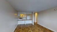 This single-floor residence showcases an open-concept layout that seamlessly connects the living area and kitchen, creating a cohesive and functional living space. The flooring throughout is warm-toned hardwood-style planks, adding a welcoming character to the interior.

The kitchen is positioned at the rear of the open space and is equipped with white cabinetry providing ample storage, white appliances including a refrigerator and range, and a sink set beneath overhead cabinets. A peninsula with a darker countertop defines the boundary between the kitchen and living area, offering additional workspace and the potential for casual bar-style seating.

The living area is generously proportioned, offering flexibility for various furniture arrangements. Neutral-toned walls complement the flooring and contribute to a bright, clean aesthetic. A textured popcorn ceiling is present throughout, and recessed lighting in the kitchen adds warm illumination to the cooking space.

A hallway visible from the main living area leads further into the unit, suggesting additional rooms beyond the open living zone. White baseboards frame the space cleanly, reinforcing the light, neutral palette throughout.