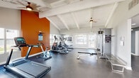Located in Denver, CO, this well-appointed fitness center offers a generously sized, single-floor exercise space designed to accommodate a full range of workout activities. The main exercise area is defined by striking vaulted ceilings with white-painted exposed beam detailing and wood paneling, creating an open and airy atmosphere. Multiple ceiling fans are mounted throughout, ensuring consistent airflow during workouts. Large windows line the walls, allowing natural light to fill the space and providing views of the surrounding exterior grounds. The rubber-floored exercise area is equipped with a variety of cardio machines, including treadmills, ellipticals, and stationary bikes, as well as a cable weight machine, a rowing machine, and a weight bench for strength training. A bold, color-saturated mural of the Denver city skyline at sunset serves as a dramatic focal point in a connected lower-ceilinged section of the space, which features recessed lighting and additional strength equipment. The fitness center's color palette — neutral gray walls offset by a vibrant orange accent wall — contributes to an energizing yet focused environment. Overall, the space is thoughtfully designed to provide a comprehensive and motivating fitness experience.