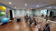This single-floor fitness studio is purpose-built for cycling and group exercise, offering a clean, energizing environment for focused workouts. The room features a row of professional-grade stationary spin bikes arranged to maximize space and allow for comfortable movement between stations. A large floor-to-ceiling mirror spans one wall, enhancing the sense of space while allowing participants to monitor their form during sessions. A wall-mounted display screen is positioned for easy viewing, ideal for streaming guided classes or workout content. The studio is finished with warm hardwood-style flooring that adds both durability and aesthetic appeal, complemented by soft sage-green walls that create a calm, motivating atmosphere. Recessed lighting throughout the ceiling provides bright, even illumination across the entire space. A plyo box is also present, indicating the room's versatility for cross-training and functional fitness beyond cycling. The layout is open and uncluttered, allowing for flexible use of the floor area. Ventilation is well-integrated into the ceiling, ensuring comfort during intense workouts. Overall, the studio offers a well-equipped, thoughtfully designed environment suited for both individual training and group fitness programming.