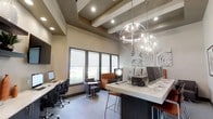 This contemporary business center is thoughtfully designed to support both focused work and collaborative tasks. The space is immediately distinguished by its striking coffered ceiling with exposed beams, which adds architectural depth and a sense of sophistication. Recessed lighting throughout ensures even, functional illumination, complemented by a bold spherical chandelier that serves as a dramatic focal point and elevates the overall aesthetic.

The layout is well-organized, offering multiple work zones to accommodate a variety of needs. A row of desktop computers is arranged along a sleek, light-colored countertop island at bar height, complete with matching stools — ideal for quick tasks or side-by-side collaboration. Along the windowed wall, additional individual workstations with ergonomic chairs provide a quieter setting for more focused work. The windows are fitted with roller shades, allowing for adjustable natural light and views to the exterior.

A comfortable seating nook with upholstered chairs and a bistro-style table offers a more relaxed environment for informal meetings or casual work. Decorative geometric wall art adds a modern, creative touch to the neutral-toned walls. The cohesive design balances functionality with a polished, professional ambiance throughout.