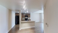 This single-floor residence offers a thoughtfully designed open-concept layout that connects the kitchen and living areas into one cohesive, airy space. The kitchen is a standout feature, equipped with rich dark-wood cabinetry, granite countertops with subtle veining, and a full suite of black appliances including a refrigerator, range, and over-the-range microwave. A peninsula counter extends from the kitchen, providing additional prep space and a natural divider between the kitchen and the living area without interrupting the open feel. Track lighting and recessed ceiling lights illuminate the kitchen and adjacent spaces efficiently. The flooring throughout is a warm-toned hardwood, adding a sense of continuity and warmth across the entire floor plan. Neutral, light-colored walls and white baseboards keep the space feeling clean and modern. The living area is generously proportioned, benefiting from natural light through a window at the far end. The smooth, textured ceiling adds a subtle architectural detail overhead. Overall, the space presents a practical yet polished interior well-suited for everyday living.