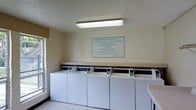 This shared laundry facility offers a practical, no-frills space designed for communal use. The room features a row of top-loading washing machines lined against a painted brick wall, accompanied by dryers positioned along the same wall. A continuous counter surface runs above and beside the machines, providing a convenient area for folding or staging laundry. Overhead, a fluorescent strip light ensures the space is well-lit throughout. A window along one wall allows for some natural light to supplement the artificial lighting. The walls are finished in painted brick, giving the room a utilitarian character. A clearly visible "Statement of Responsibility" sign is posted on the wall, outlining usage rules such as prohibitions on overloading, dyeing, using flammable items, and leaving unattended clothes. The flooring is a light-toned material that complements the neutral palette of the room. The overall ambiance is functional and straightforward, designed to accommodate the laundry needs of multiple users efficiently. The space maintains a clean, organized layout that maximizes accessibility to all machines.