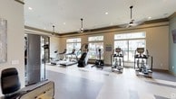 Located at 22 Campbell Scenic Dr in Hattiesburg, Mississippi, this single-floor fitness center offers a comprehensive workout facility designed to support a full range of exercise routines. The space is thoughtfully divided into distinct workout zones, each finished with a polished, upscale aesthetic characterized by crown molding, recessed lighting, pendant ceiling fans, and a warm neutral-and-blue color palette.

The cardio area is bright and open, lined with large windows that invite abundant natural light and offer views of the surrounding outdoor landscape including palm trees and a pool area. This section houses multiple treadmills, elliptical trainers, and stair climbers, all arranged to take advantage of the scenic views.

A dedicated strength and free weight zone features a striped performance flooring area, a full rack of dumbbells, a weight bench, and a multi-function strength machine. The muted blue accent walls and pendant lighting give this area a focused, calm atmosphere.

A separate functional training area contains cable machines, a rowing machine, and additional strength equipment, complemented by tasteful wall art that adds a refined touch. Wall-mounted televisions throughout the space provide entertainment during workouts.

Together, these zones create a fully equipped, visually cohesive fitness facility that balances functionality with an elevated, resort-style design sensibility.