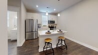 This two-floor space is thoughtfully designed with a cohesive aesthetic that balances modern finishes with warm, inviting details. The lower level serves as the main living and entertaining area, anchored by rich, dark hardwood flooring that runs throughout. The open-concept layout connects a spacious living room to a fully equipped kitchen, creating a seamless flow between the two areas. Recessed lighting illuminates the living room, which features neutral-toned walls, clean white baseboards, and a structural beam detail that adds architectural character to the ceiling. A window allows natural light to filter in alongside the well-placed recessed fixtures.

The kitchen is a functional highlight, outfitted with white shaker-style cabinetry, stainless steel appliances including a French door refrigerator, microwave, and range, as well as a peninsula countertop with seating for two. Pendant lighting adds a polished touch above the counter.

The upper level includes a well-sized bedroom with plush carpeting, a ceiling fan, and a window fitted with blinds for light control. The bathroom on this level is clean and bright, featuring a white vanity with drawers, a framed mirror, a tub, and dark hardwood-style flooring that ties back to the lower level's design language.