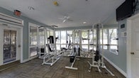 Located at 10725 Abercorn Ext in Savannah, Georgia, this fitness center offers a well-equipped, single-floor workout space designed to support a comprehensive strength training routine. The room is lined with large windows on multiple walls, flooding the interior with abundant natural light and providing pleasant views of the surrounding outdoor greenery. The light-toned walls in a soft blue-gray palette contribute to an open and refreshing atmosphere, complemented by patterned carpet tile flooring that adds both texture and durability underfoot. A ceiling fan is centrally mounted, ensuring comfortable air circulation throughout the space during workouts. The room is outfitted with multiple white cable and weight machines, including pull-down stations, a multi-functional cable crossover unit, and various resistance training apparatuses, accommodating a range of upper and lower body exercises. Recessed lighting supplements the natural light for consistent illumination at all hours. The textured ceiling adds a classic finish to the space. Overall, the fitness center presents a bright, functional, and well-maintained environment suited for residents or members seeking a convenient and adequately equipped place to exercise.