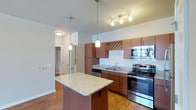 This two-story residence is thoughtfully designed to balance functionality and comfort across its multiple levels. The main floor centers around an open-concept kitchen and living area, where warm hardwood-style flooring flows throughout. The kitchen is equipped with rich wood cabinetry, granite countertops, stainless steel appliances including a range and microwave, a double sink, and a central island with pendant lighting overhead. A decorative wine rack accent adds character above the upper cabinets, while track lighting illuminates the workspace effectively.

The living area benefits from soaring vaulted ceilings that create an airy, expansive feel, complemented by a front entry door with transom windows that invite natural light into the space. The upper level features an open loft-style area with a ceiling fan, hardwood-style flooring, and a staircase railing detail, offering a versatile secondary living space.

The bedroom is carpeted for comfort and includes a ceiling fan, with convenient access to both a walk-in closet and an en-suite bathroom. Two bathrooms are present in the residence, each featuring granite-topped wood vanities and tub/shower combinations with tile or panel surrounds. Clean neutral tones and consistent finishes throughout lend the space a cohesive, well-appointed character.