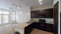 This 1-bedroom, 1-bathroom unit in Houston, TX (77025) is a single-story residence thoughtfully designed for comfortable urban living. The open-concept kitchen serves as the centerpiece of the living area, outfitted with rich dark-espresso cabinetry, granite countertops, and a full suite of black appliances including a range, microwave, dishwasher, and refrigerator with an ice and water dispenser. A peninsula with an integrated sink extends the counter space and creates a natural divide between the kitchen and living area, while pendant lighting and track lighting illuminate the space stylishly. Wood-style flooring runs throughout the kitchen and entryway, adding warmth and easy maintenance. The bedroom is carpeted and benefits from large windows that allow natural light to filter in. The bathroom is well-appointed with tile flooring, a soaking tub with tiled surround, a dark-cabinetry vanity with a granite countertop, and direct access to a walk-in closet complete with built-in shelving. The overall design palette is neutral and cohesive, combining clean white walls with dark fixtures and granite surfaces for a modern, polished aesthetic throughout.