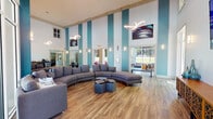 Located at 1560 San Luis Rd in Tallahassee, Florida, this community clubhouse is a well-appointed gathering and amenity space designed with a bold, contemporary aesthetic. The centerpiece of the facility is a soaring double-height lounge area accented by teal and white vertical striping on the walls, warm wood-look flooring, and sculptural pendant lighting that adds a modern artistic flair. Large floor-to-ceiling windows and sliding glass doors flood the interior with abundant natural light while offering direct views of the outdoor pool area and surrounding greenery. The lounge is furnished with curved sectional sofas, accent chairs, and geometric coffee tables, creating an inviting and social atmosphere. Vibrant large-scale artwork adorns the walls throughout, adding color and energy to the space. A dedicated media area features a wall-mounted flat-screen television above a decorative credenza, framed by expansive windows that overlook the landscaped exterior. The fitness center is generously sized and well-lit, equipped with a comprehensive selection of cardio machines, cable systems, free weights, and weight benches, all set on matching wood-look flooring with mirrored walls and large windows providing views of the outdoors. The cohesive teal color palette ties all areas of the clubhouse together into a unified, resort-style environment.