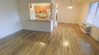 This single-floor space offers an open-concept layout that seamlessly connects the living area and kitchen, creating a spacious and functional environment. Gleaming hardwood floors extend throughout, adding warmth and a polished aesthetic to the interior.

The kitchen is defined by a raised breakfast bar peninsula that separates it from the living area while maintaining an open feel. It features honey-toned wooden cabinetry, a white appliance suite including a range and microwave, and a built-in sink. The pass-through design enhances communication between the kitchen and the adjoining living space.

The living area is generously proportioned, benefiting from the continuity of the hardwood flooring and neutral gray wall tones that contribute to a calm, understated ambiance. A decorative pendant light fixture hangs overhead, providing a focal point and adding a touch of character to the space.

A hallway visible toward the rear of the unit leads to additional rooms, suggesting a practical and well-organized floor plan. Natural light filters into the space, further enhancing the open and airy atmosphere throughout this thoughtfully designed single-level home.