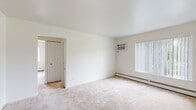 This single-floor residential unit offers a clean, neutral aesthetic throughout, with white walls and light-toned carpeting creating a bright and airy atmosphere across the living spaces. The generously sized main living area is a standout feature, offering ample room for a variety of furniture arrangements. Large windows dressed with vertical blinds invite natural light and provide views of surrounding greenery, while a wall-mounted air conditioning unit and baseboard heating ensure year-round comfort. A closet with classic bi-fold doors adds practical storage to the main living space. The bedroom is well-proportioned, featuring its own window with vertical blinds, baseboard heating, and a ceiling fan with a light fixture, contributing to both comfort and functionality. The kitchen is outfitted with warm oak cabinetry, white laminate countertops, a stainless steel sink, and white appliances including an electric range and dishwasher, all set atop durable tile flooring. The full bathroom includes a tub-shower combination, a toilet, and a vanity with a granite-patterned countertop and a large mirror above, illuminated by a multi-bulb light bar. The overall layout is efficient and functional, offering a comfortable living environment with consistent finishes throughout.