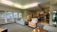 This single-story space is thoughtfully designed to offer a comfortable and inviting atmosphere, combining a well-appointed lounge area with a stylish bar section. The lounge features a tray ceiling with integrated ambient lighting that casts a warm, even glow throughout the room, enhancing its refined character. Neutral green-gray walls and plush, light-toned upholstered furniture — including a sofa and accent armchair — create a relaxed yet polished seating area anchored by a carpeted floor. Natural light filters in through slatted blinds on the windows, complementing the warm artificial lighting. A wooden side table with a decorative lamp adds a homey touch to the seating arrangement. Toward the back of the space, the flooring transitions to hardwood as the area opens into a bar section, complete with bar stools, pendant track lighting overhead, and a well-stocked display shelf behind the counter. This combination of textures, lighting layers, and functional zones gives the space a welcoming, upscale communal feel suited for socializing and relaxation.