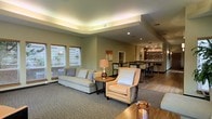 This single-story space is thoughtfully designed to offer a comfortable and inviting atmosphere, combining a well-appointed lounge area with a stylish bar section. The lounge features a tray ceiling with integrated ambient lighting that casts a warm, even glow throughout the room, enhancing its refined character. Neutral green-gray walls and plush, light-toned upholstered furniture — including a sofa and accent armchair — create a relaxed yet polished seating area anchored by a carpeted floor. Natural light filters in through slatted blinds on the windows, complementing the warm artificial lighting. A wooden side table with a decorative lamp adds a homey touch to the seating arrangement. Toward the back of the space, the flooring transitions to hardwood as the area opens into a bar section, complete with bar stools, pendant track lighting overhead, and a well-stocked display shelf behind the counter. This combination of textures, lighting layers, and functional zones gives the space a welcoming, upscale communal feel suited for socializing and relaxation.
