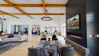 This expansive single-floor clubhouse is designed with a contemporary farmhouse aesthetic, combining warm wood accents with crisp white walls and light hardwood flooring throughout. The centerpiece of the main lounge is a striking floor-to-ceiling dark shiplap fireplace wall, topped with a large-scale landscape painting and anchored by a linear gas fireplace with a concrete hearth. Soaring ceilings with exposed natural wood beams and coffered detailing add architectural drama, while oversized windows and glass French doors flood the space with natural light and offer views of the outdoor amenity area. A curated seating arrangement of leather and upholstered chairs surrounds layered area rugs, creating an inviting gathering atmosphere. A dedicated coworking zone features individual workstations with task chairs, ideal for residents working remotely. Adjacent game and lounge areas include a pool table, ping pong table, and multiple seating clusters anchored by a geometric chandelier and a dark shiplap accent wall with a mounted television. The kitchen and bar area is outfitted with a long counter, bar seating, open shelving, and pendant lighting, providing a functional space for casual dining and entertaining. The seamless open floor plan connects each zone fluidly, making the clubhouse a versatile and welcoming community hub.