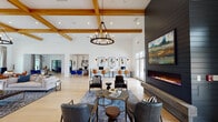 This expansive single-floor clubhouse is designed with a contemporary farmhouse aesthetic, combining warm wood accents with crisp white walls and light hardwood flooring throughout. The centerpiece of the main lounge is a striking floor-to-ceiling dark shiplap fireplace wall, topped with a large-scale landscape painting and anchored by a linear gas fireplace with a concrete hearth. Soaring ceilings with exposed natural wood beams and coffered detailing add architectural drama, while oversized windows and glass French doors flood the space with natural light and offer views of the outdoor amenity area. A curated seating arrangement of leather and upholstered chairs surrounds layered area rugs, creating an inviting gathering atmosphere. A dedicated coworking zone features individual workstations with task chairs, ideal for residents working remotely. Adjacent game and lounge areas include a pool table, ping pong table, and multiple seating clusters anchored by a geometric chandelier and a dark shiplap accent wall with a mounted television. The kitchen and bar area is outfitted with a long counter, bar seating, open shelving, and pendant lighting, providing a functional space for casual dining and entertaining. The seamless open floor plan connects each zone fluidly, making the clubhouse a versatile and welcoming community hub.
