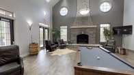 Located in Brighton 3050, this single-level communal space is designed as an upscale social and recreation lounge, blending modern and rustic design elements throughout. The room commands immediate attention with its dramatically vaulted ceilings, which soar overhead and are punctuated by circular porthole windows that flood the interior with natural light. Wood-look flooring runs throughout, grounding the space with a warm, earthy tone that complements the cool grey walls.

The centrepiece of the space is a floor-to-ceiling stacked stone fireplace, its substantial chimney rising impressively against the feature wall, creating a striking focal point. Adjacent to the fireplace, a sitting area features leather armchairs arranged on a cowhide rug, offering an intimate and comfortable retreat. A full-size billiards table occupies the central zone, serving as the social hub of the room.

A generous lounge area is outfitted with a large sectional sofa and wall-mounted flat-screen televisions, ideal for relaxed gatherings. A wet bar with counter seating and pendant lighting adds further functionality. Rustic barrel accents and curated wall art reinforce the space's refined yet relaxed character. Black-framed glass doors provide direct outdoor access, enhancing the sense of openness and connection to the surrounding environment.