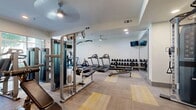 Located at 1700 W Frankford Rd in Carrollton, Texas, this fitness center offers a well-appointed, single-floor workout facility designed to support a full range of exercise routines. The space features a modern, clean aesthetic with neutral-toned walls, recessed and ceiling fan lighting, and a patterned carpet floor accented with yellow geometric detailing that adds visual interest throughout.

The cardio section is equipped with multiple Matrix treadmills and a stationary bike, positioned to maximize the open layout. A dedicated strength training area houses a multi-functional cable machine and weight rack, with a full set of dumbbells neatly arranged for easy access. An adjustable weight bench and additional free-weight equipment round out the strength training offerings. A corner of the space is dedicated to functional fitness, featuring stability balls and yoga accessories stored alongside a compact beverage refrigerator.

Large floor-to-ceiling windows on two walls flood the interior with abundant natural light and provide views of the surrounding outdoor greenery, creating an inviting and energizing atmosphere. A wall of mirrors further enhances the sense of space and openness. A mounted flat-screen TV adds entertainment options for users during workouts. The facility is accessible via glass-paneled entry doors and is equipped with clearly marked exit signage, ensuring a safe and functional environment.