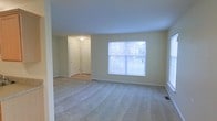 This single-story space features a generously sized main room finished with plush, neutral-toned wall-to-wall carpeting that adds warmth and comfort underfoot. The walls are painted in a soft, light cream tone, contributing to a clean and airy atmosphere throughout. A large window along one wall allows natural light to fill the room, offering a view of the surrounding exterior greenery. The white trim and baseboards provide a crisp contrast against the light walls, lending the space a polished, well-maintained appearance. An adjacent hallway is visible from the main room, featuring a flush-mount ceiling light fixture and two white paneled doors, likely leading to closets or additional rooms, adding functional storage potential. Vinyl or hardwood-style flooring is visible in the hallway, distinguishing it from the carpeted main area. Subtle details such as floor vents indicate a central HVAC system. Overall, the space presents a straightforward, move-in-ready layout with a neutral palette well-suited for a variety of furnishing styles and uses.