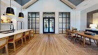 Located at 3 Whisperwood Way in Candler, NC, this single-story space offers a thoughtfully designed open-concept layout that maximizes both function and aesthetic appeal. The interior is anchored by dramatic vaulted ceilings with exposed wood beam detailing, lending a sense of architectural grandeur throughout. Wide-plank hardwood flooring runs continuously across the space, unifying each zone with warmth and cohesion.

The living room is a standout feature, divided into distinct yet connected seating areas. One lounge area showcases a statement accent wall in muted blue-gray, adorned with a curated display of decorative oars — a distinctive artistic touch. Comfortable, neutral-toned furnishings and layered textures create an inviting, relaxed atmosphere. A second seating nook benefits from floor-to-ceiling windows that flood the space with abundant natural light and frame views of the surroundings.

Elegant steel-framed glass partition doors add a modern industrial contrast to the otherwise soft, organic palette. The open floor plan also accommodates dining areas, seamlessly integrated into the overall flow. The interplay of high ceilings, natural light, warm wood tones, and carefully chosen furnishings gives this Candler property a refined yet welcoming character throughout.