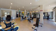 This fitness center is thoughtfully designed to support a comprehensive range of workout routines within a single, open-plan floor. The space features light-toned herringbone-patterned flooring throughout, recessed lighting, and soft blue and neutral wall tones that create a clean, energizing atmosphere.

The cardio zone is lined with a row of True-brand treadmills and elliptical machines, positioned to face large glass double doors that allow natural light to flood the space and offer views of the surrounding outdoor area. A wall clock and full-length mirrors enhance the functionality of this section.

Adjacent to the cardio area, a dedicated strength training zone houses a variety of selectorized resistance machines and cable equipment, supported by structural columns that define the area without enclosing it. A spin bike and additional cardio equipment are also present in this zone.

A flexibility and functional fitness area includes stability balls in multiple sizes mounted on the wall, medicine balls, a yoga/exercise mat display, and ample open floor space for stretching or bodyweight training.

The free weights section offers a well-organized rack of dumbbells and kettlebells, a wall-mounted flat-screen television, and a large framed mirror. Completing the amenity, a pair of massage chairs and a water bottle filling station provide post-workout recovery options, making this a fully self-contained wellness facility.